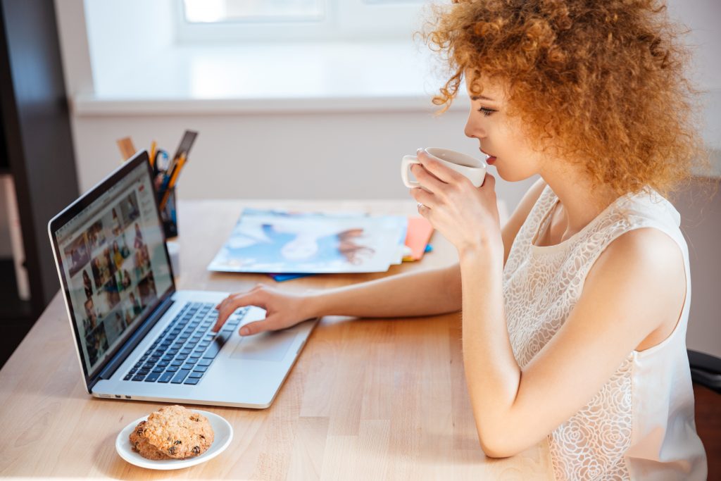 Profile of attractive charming curly young woman photographer drinking coffee and working with laptop on workplace